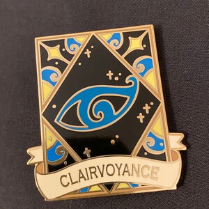 May include: Gold-toned enamel pin with a black background and a blue eye design. The pin features a white banner with the text "Clairvoyance".