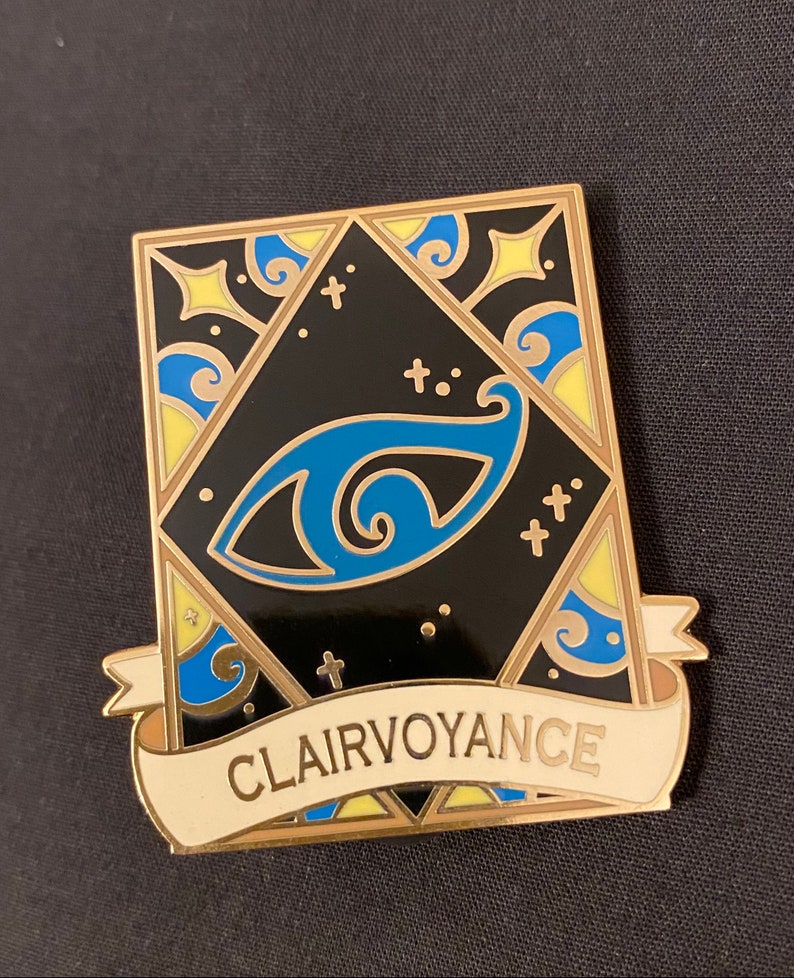 May include: Gold-toned enamel pin with a black background and a blue eye design. The pin features a white banner with the text "Clairvoyance".