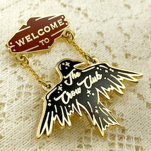 May include: A gold-toned metal enamel pin featuring a maroon banner with the text WELCOME TO connected by two gold chains to a black crow-shaped pendant with the text The Crow Club and small star accents in a gold-toned metal finish.