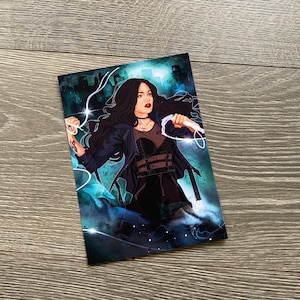 May include: A woman with long black hair is standing in front of a city skyline. She is wearing a black leather jacket, a black top, and black pants. She is holding a glowing white object in her right hand. The background is a dark blue and green color with white stars.