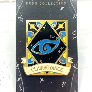 May include: A gold and blue enamel pin with a black background. The pin features a stylized eye with a blue iris and a gold pupil. The pin is surrounded by a gold frame with a yellow and blue design. The pin has a white banner with the word "Clairvoyance" in gold letters. The pin is on a black card with the words "Shadowhunters Rune Collection" and "Designed by @azura.arts".