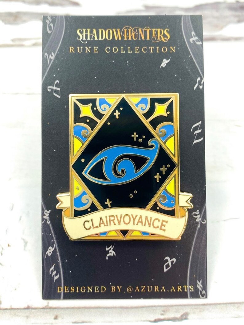May include: A gold and blue enamel pin with a black background. The pin features a stylized eye with a blue iris and a gold pupil. The pin is surrounded by a gold frame with a yellow and blue design. The pin has a white banner with the word "Clairvoyance" in gold letters. The pin is on a black card with the words "Shadowhunters Rune Collection" and "Designed by @azura.arts".