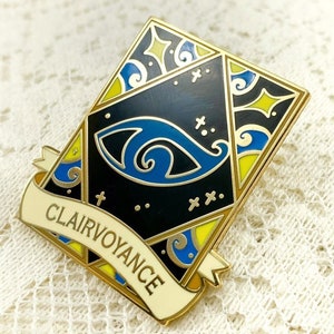 May include: Gold-toned enamel pin with a black background and a blue eye design. The pin features a yellow and blue border with a white banner that reads "Clairvoyance".