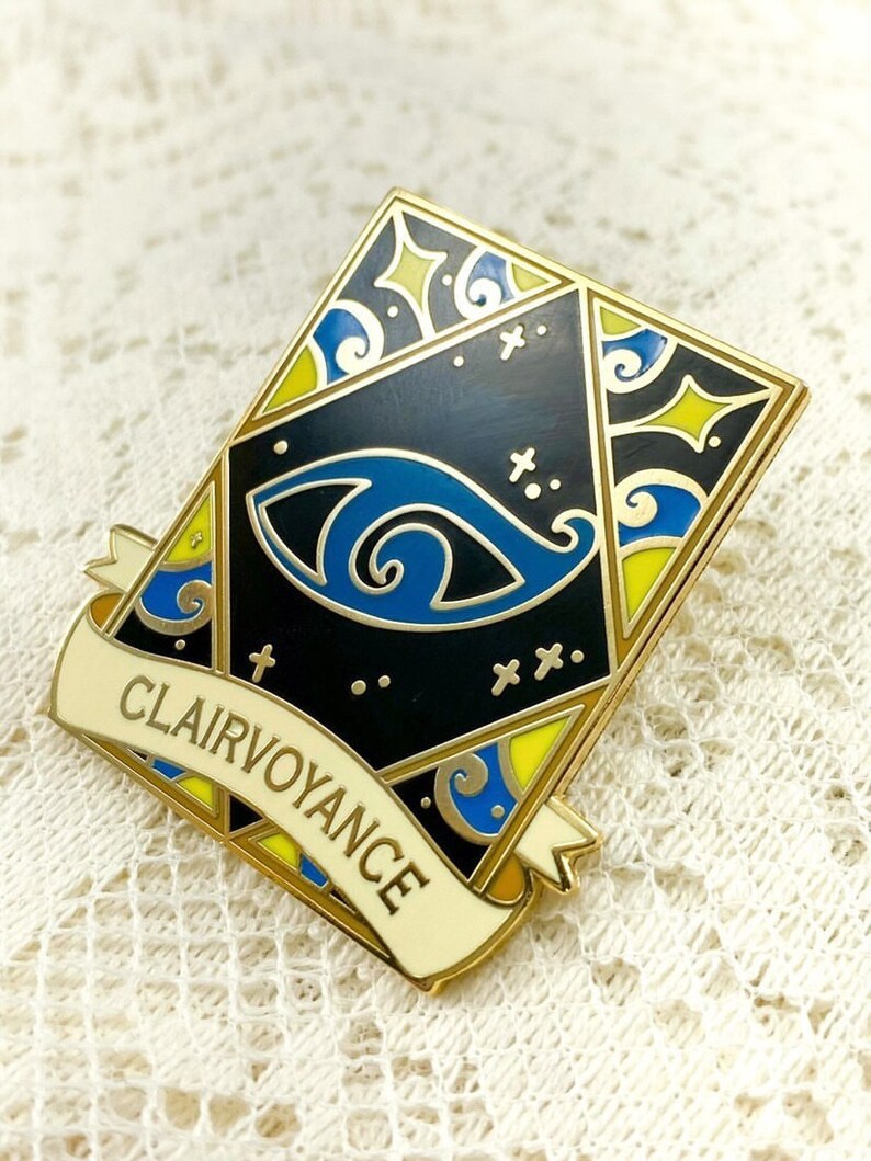 May include: Gold-toned enamel pin with a black background and a blue eye design. The pin features a yellow and blue border with a white banner that reads "Clairvoyance".
