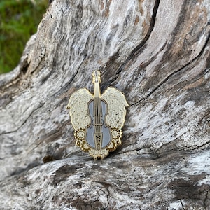 May include: Gold-tone metal pin featuring a silver violin with wings and floral accents.