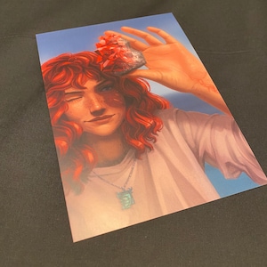 May include: Art print featuring a person with red curly hair winking, holding a red crystal against a blue sky background. The person wears a white shirt and a necklace with a green pendant. The artwork has a soft, painterly style.