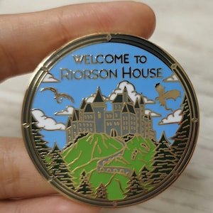 May include: A gold-colored enamel pin with a blue background and the text "Welcome to Riorson House". The pin features a gray castle on a green hill with a winding road leading up to it. Two blue dragons are flying in the sky above the castle.
