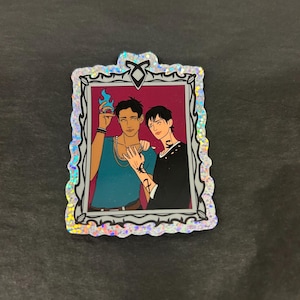 May include: A rectangular sticker featuring an illustration of two men. One man is wearing a teal shirt and holding a blue flame. The other man is wearing a black shirt. The sticker has a glittery, silver border.