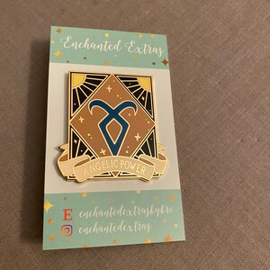 Könnte beinhalten: Goldene und braune Emaille-Anstecknadel mit einem blauen Symbol und dem Schriftzug "Angelic Power" auf einem Banner. Die Anstecknadel befindet sich auf einer Karte mit dem Text "Enchanted Extras" und Social-Media-Handles.