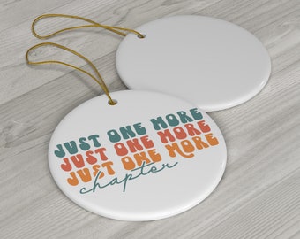 Puede incluir: Adorno de cerámica blanca con una cuerda dorada para colgar. El adorno tiene el texto "Just One More Just One More Just One More chapter" en una fuente retro con colores naranja, verde azulado y rojo.