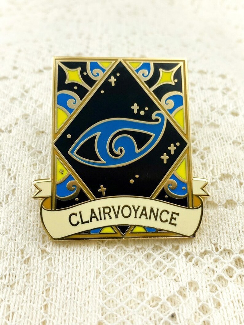 May include: Gold-toned enamel pin with a blue eye design and a banner that reads "Clairvoyance". The eye is surrounded by a black background with yellow and blue swirls.
