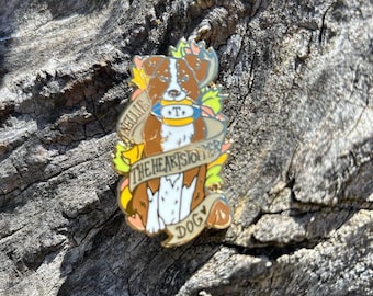 May include: A gold enamel pin featuring a brown and white dog with a blue and yellow collar. The dog is surrounded by flowers and a banner that reads "Nellie the Heart Stomper Dog".