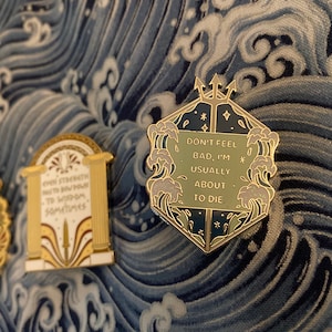 Pode incluir: Três pinos de esmalte dourado com designs diferentes. O primeiro pino apresenta um pergaminho com o texto "Mesmo a força precisa se curvar à sabedoria, às vezes". O segundo pino apresenta uma coluna com uma espada e o texto "*P*0". O terceiro pino apresenta um design hexagonal azul escuro e verde com um tridente e o texto "Não se sinta mal, geralmente estou prestes a morrer".