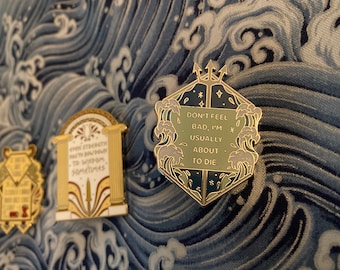 Puede incluir: Tres pines de esmalte dorado con diferentes diseños. El primer pin presenta un rollo con el texto "Incluso la fuerza necesita inclinarse ante la sabiduría, a veces". El segundo pin presenta una columna con una espada y el texto "*P*0". El tercer pin presenta un diseño hexagonal azul oscuro y verde con un tridente y el texto "No te sientas mal, normalmente estoy a punto de morir".