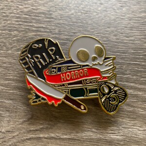 Pode incluir: Pin de esmalte dourado com uma caveira, uma lápide com o texto "R.I.P." e uma faca com cabo vermelho. A caveira segura uma faixa vermelha com o texto "HORROR".