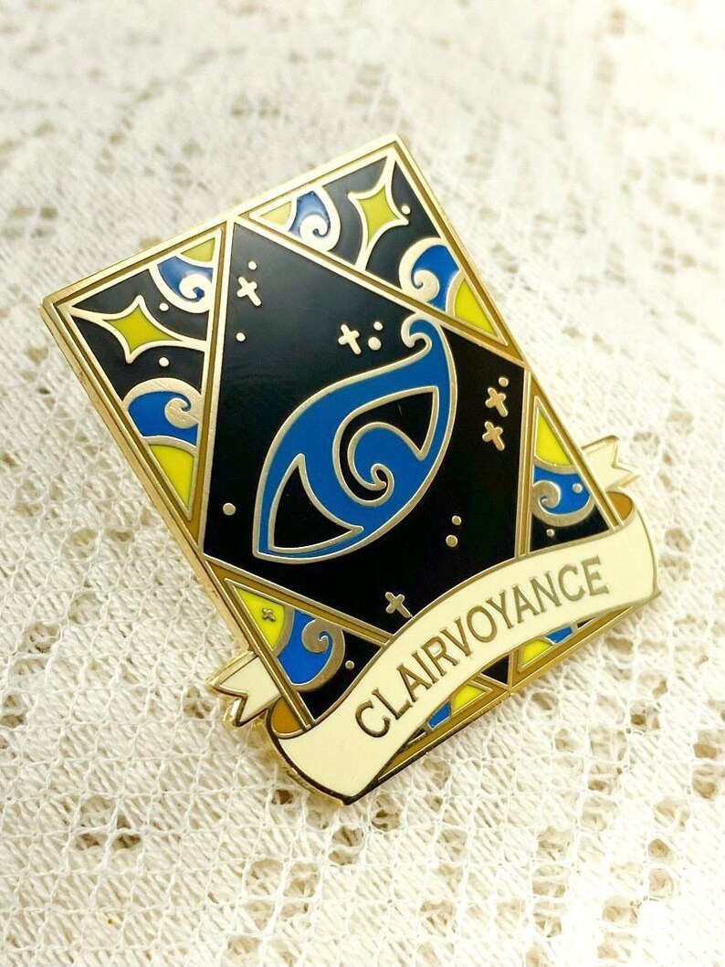 May include: Gold-toned enamel pin with a black background and a blue eye design. The pin features a white banner with the text "Clairvoyance" in yellow lettering.