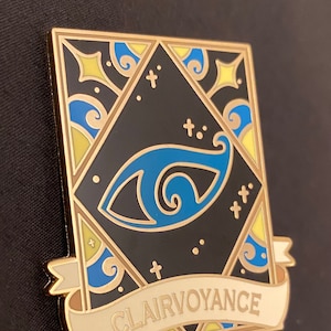 May include: Gold-toned enamel pin with a blue eye design and a banner that reads "Clairvoyance". The eye is surrounded by a geometric pattern with yellow and blue accents.