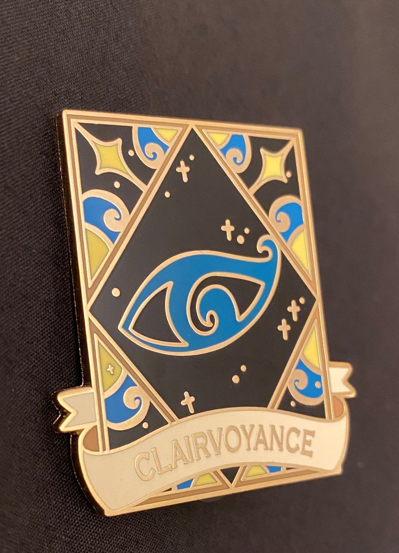May include: Gold-toned enamel pin with a blue eye design and a banner that reads "Clairvoyance". The eye is surrounded by a geometric pattern with yellow and blue accents.