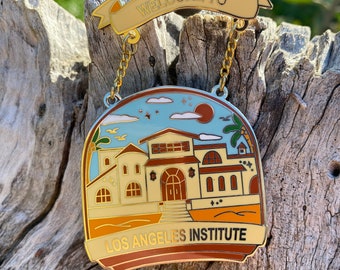 May include: Gold enamel pin with a banner that reads "Welcome to Los Angeles Institute". The pin depicts a building with a red roof and a palm tree in front of it. The pin is hanging from a gold chain.