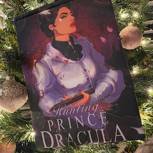 Può includere: Una copertina di libro con una donna in una camicia bianca con macchie di sangue, che tiene un paletto di legno. Il testo sulla copertina recita "Hunting Prince Dracula".