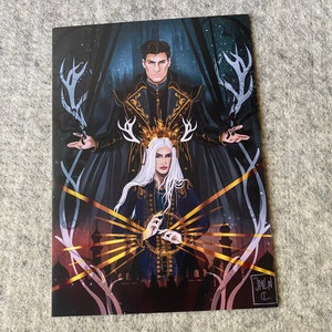 Peut inclure: Une impression d'art fantastique représentant deux personnages, l'un vêtu d'une robe noire et dorée et l'autre d'une robe bleue et dorée. Les personnages sont entourés de bois de cerf et d'une lueur dorée. L'impression est signée "JALA" en bas à droite.