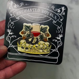 May include: Enamel pin featuring a gold crown, a wine glass with red liquid, and a black snake. The pin is set against a black backdrop with the words "Enchanted Royals" in white. The pin is on a black card.