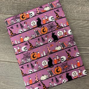 May include: Purple fabric with a Halloween-themed pattern. The design includes black cats, witch hats, pumpkins, potion bottles, skulls, and spiderwebs. The fabric is suitable for crafting projects.