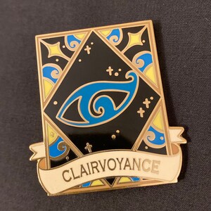 May include: Gold-toned enamel pin with a black background and a blue eye design. The pin has a white banner with the word "Clairvoyance" written in gold lettering.
