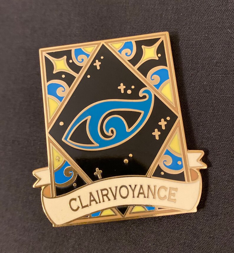 May include: Gold-toned enamel pin with a black background and a blue eye design. The pin has a white banner with the word "Clairvoyance" written in gold lettering.
