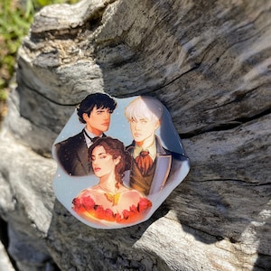 May include: A sticker featuring three characters from a fantasy novel. The characters are a man with dark hair, a man with white hair, and a woman with brown hair. The woman is wearing a red dress with a gold butterfly brooch. The sticker is on a piece of weathered wood.