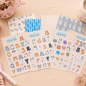 May include: Four sheets of stickers featuring cat-themed alphabet letters, numbers, and small icons. Each sheet has a grid background and the text Cleverly Printed. The stickers include various cat illustrations in orange, gray, and white colors.