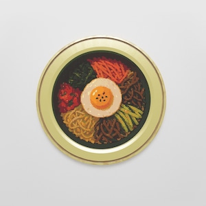 Op de afbeelding: Een rond, ingelijst kunstwerk dat een gepixeld kommetje bibimbap, een Koreaans gemengd rijstgerecht, afbeeldt. Het gerecht bevat een gebakken ei, groenten en rijst, met een donkergroene kom en een goudkleurige lijst.