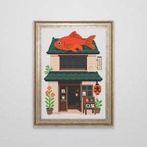 Könnte beinhalten: Gerahmtes Kreuzstich-Kunstwerk eines japanischen Geschäfts. Das Gebäude ist beige mit einem grünen Dach und einem großen orangefarbenen Fisch oben. Eine rote Laterne hängt in der Nähe des Eingangs. Topfpflanzen und eine Holzauslage ergänzen die Szene.