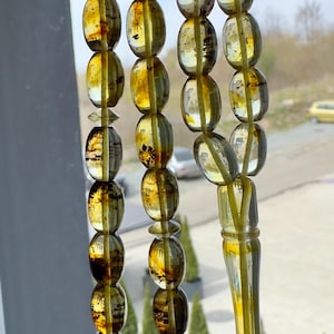 Puede incluir: Un collar de cuentas de oración de color ámbar con cuentas de forma ovalada. Las cuentas tienen una calidad translúcida con variaciones de color, que van del amarillo claro al marrón oscuro. Las cuentas están ensartadas con un hilo amarillo a juego.