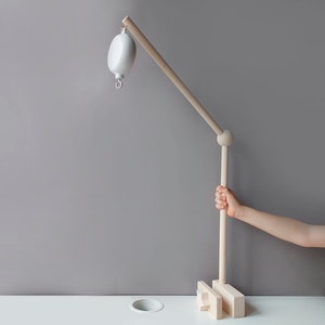 May include: A wooden baby mobile arm with a white ball hanging from it. The arm is attached to a wooden base with a hand holding the arm.