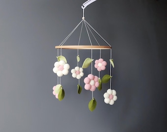 May include: A baby mobile featuring felt flowers and leaves in shades of pink, white, and green. The flowers are spherical and the leaves are teardrop-shaped. The mobile hangs from a wooden ring and is suspended by white strings.