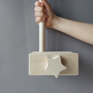 May include: A wooden toy hammer with a star-shaped head and a wooden handle. The hammer is painted in a natural wood color.
