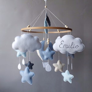 May include: A baby mobile with a wooden ring, blue and white clouds, stars, and a hot air balloon. The cloud on the right has the name "Emilio" embroidered on it.