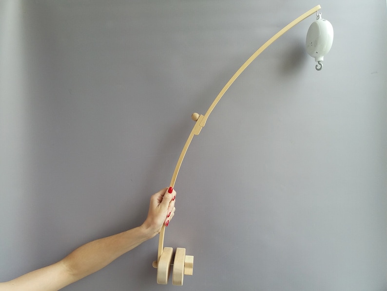 K&ouml;nnte beinhalten: Ein Holzarm f&uuml;r ein Baby-Mobile mit einem wei&szlig;en Mobile, das daran h&auml;ngt. Der Arm ist gebogen und hat einen Holzgriff am Ende. Der Arm wird von einer Hand gehalten.