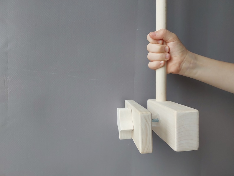 May include: A white wooden dowel rod with a knob on one end and a rectangular wooden block attached to the other end. The rod is attached to the block with a screw. The block has a smaller rectangular block attached to it with a screw.