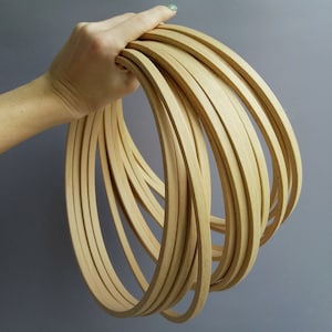Op de afbeelding: Een stapel van 10 natuurlijke houten hoepels, elk ongeveer 1,25 cm breed. De hoepels zijn in een cirkelvormige opstelling gerangschikt en worden in de hand van een persoon gehouden.