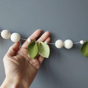 May include: A decorative garland featuring felt leaves and white pom-poms strung on a white cord. The leaves are light green, and the pom-poms are a soft white. Small wooden beads are interspersed between the leaves and pom-poms. The garland is displayed against a gray background.
