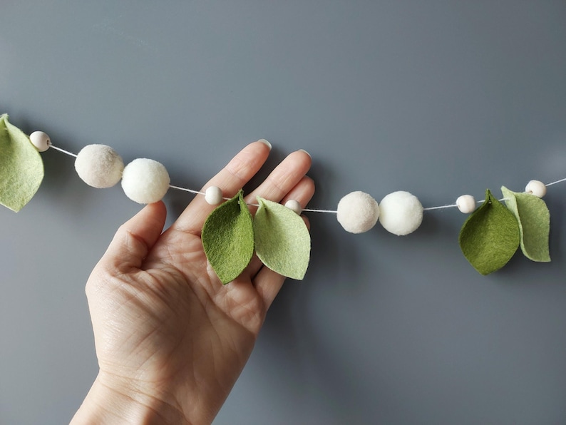 May include: A decorative garland featuring felt leaves and white pom-poms strung on a white cord. The leaves are light green, and the pom-poms are a soft white. Small wooden beads are interspersed between the leaves and pom-poms. The garland is displayed against a gray background.