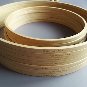 Op de afbeelding: Een set van zes houten borduurringen, allemaal met het label "Nurge No 6". De ringen zijn op elkaar gestapeld, met de grootste ring onderaan en de kleinste ring bovenaan.