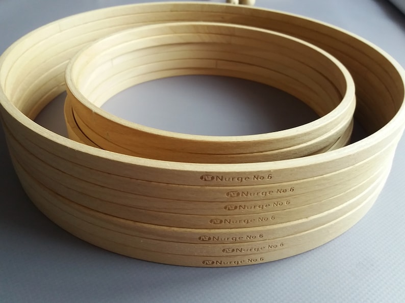 Op de afbeelding: Een set van zes houten borduurringen, allemaal met het label "Nurge No 6". De ringen zijn op elkaar gestapeld, met de grootste ring onderaan en de kleinste ring bovenaan.