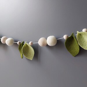 May include: A decorative garland featuring white felt balls, small wooden beads, and green felt leaves strung on a white cord. The garland is against a gray background, creating a natural, organic aesthetic.