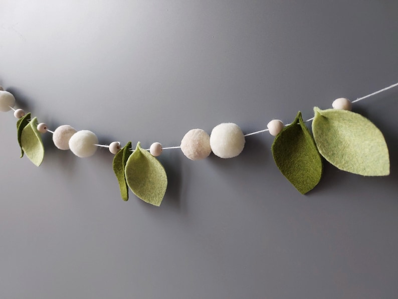 May include: A decorative garland featuring white felt balls, small wooden beads, and green felt leaves strung on a white cord. The garland is against a gray background, creating a natural, organic aesthetic.