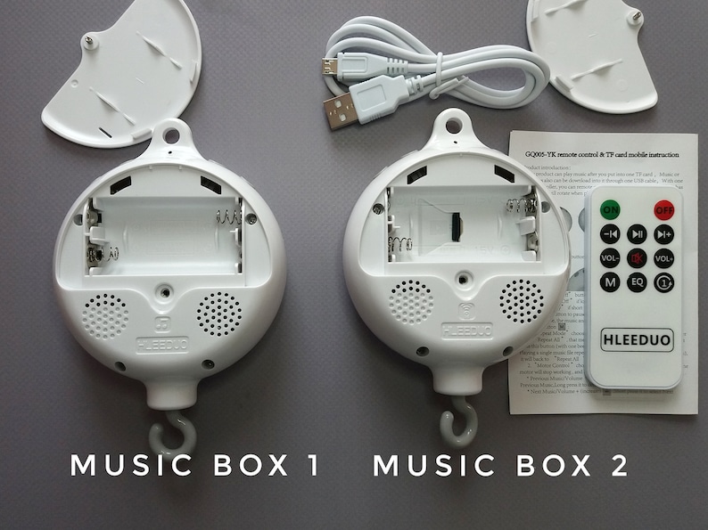 May include: Two white music boxes with open compartments revealing battery slots and a TF card slot. The music boxes have a hook for hanging and a speaker on the bottom. A white remote control with buttons for on/off, volume, play/pause, and other functions is included. The text on the remote control reads "HLEEDUO". The text on the instruction sheet reads "GQ005-TF remote control & TF card mobile instruction".