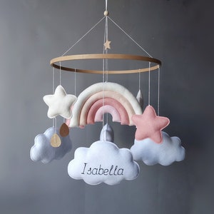 May include: A wooden baby mobile with a rainbow, clouds, and stars. The clouds are white with a light blue trim and one cloud has the name "Isabella" embroidered on it.