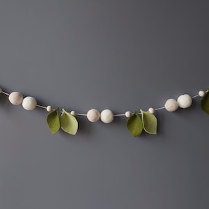 May include: A decorative garland featuring felt leaves and round, light-colored beads. The leaves are a soft green, and the beads are a mix of white and beige. The garland is strung on a thin white cord, perfect for home decor.